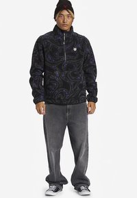 Fleecegenser i svart med lilla virvelmønster; har halvglidelås og elastiske mansjetter. Kombinert med grå vide jeans og joggesko.