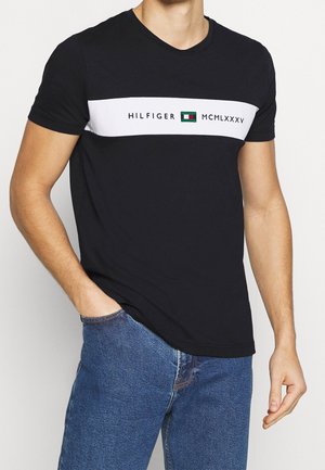 Man wearing short-sleeve black T-shirt with white horizontal stripe and text "HILFIGER MCMLXXXV," paired with blue jeans, hand in pocket.