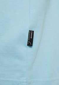 Helles blaues T-Shirt aus glattem Baumwollmaterial, mit einem seitlichen Etikett in Schwarz und Akzentnähten nahe dem Saum.