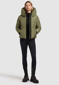 Olijf groene pufferjack met een hoge kraag, ritsluiting en een oversized capuchon. Gepaard met zwarte leggings en kniehoge laarzen. Zachte textuur.