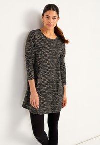 Kvinna med mörkt hår i hästsvans iförd en långärmad svart klänning med beige rektangulära mönster och svarta leggins mot en vit vägg.