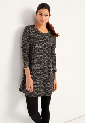 Woman with dark hair in a ponytail wearing a long-sleeve black dress with beige rectangular patterns and black leggings against a white wall.