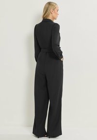 Mono largo negro de manga larga con cintura ajustada, diseño de pierna ancha y bolsillos laterales. Presenta un cuello con solapa y una textura suave.