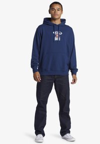 Sudadera con capucha de color azul marino con bolsillo frontal y logo blanco. Combinada con jeans de denim oscuro y zapatillas blancas.