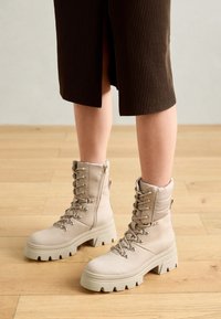 Botas de tobillo beiges con un collar acolchado, cordones y suela de goma gruesa con profundas estrías. Textura suave con acentos metálicos.