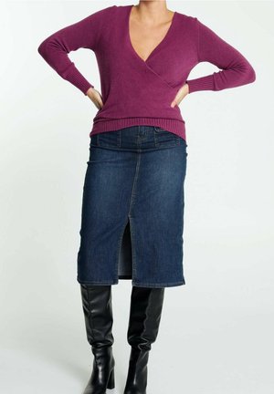 Femme portant un pull violet à col en V, une jupe en denim foncé longueur genou avec une fente à l'avant, et des bottes noires hautes, debout les mains sur les hanches.