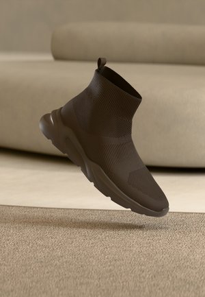 Mørkebrun sokke-stil sneakers med strikket overdel og chunky sål, ophængt over et beige tæppe i et rum med neutrale toner.