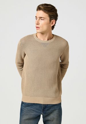 Beżowy sweter robiony na drutach z fakturowanym wzorem, okrągłym dekoltem i ściągaczami. W połączeniu z niebieskimi dżinsami, model stoi z rękami za plecami.