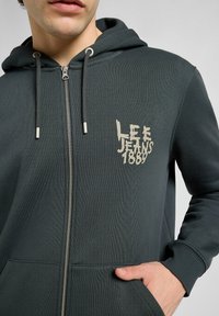Felpa con zip verde scuro con un finish testurizzato, caratterizzata da un logo frontale "LEE JEANS 1889" in beige, cordini e tasche laterali.