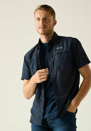 Hombre que lleva una camisa de manga corta azul oscuro sobre una camiseta azul marino y vaqueros azules, con una mano cerca del pecho y la otra en el bolsillo.