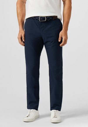 Man draagt marineblauwe broek, witte sneakers, een wit overhemd en een donker gevlochten riem tegen een effen lichte achtergrond.
