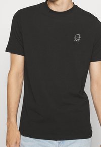 T-shirt nera in cotone con maniche corte, caratterizzata da un piccolo design grafico bianco del profilo di una persona sulla parte sinistra del petto.
