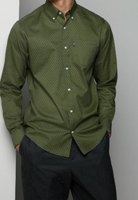 Green button-up shirt with a geometric pattern, long sleeves, and white buttons. Paired with dark pants, featuring a classic collar design.