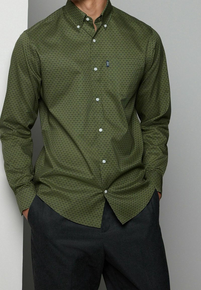Green button-up shirt with a geometric pattern, long sleeves, and white buttons. Paired with dark pants, featuring a classic collar design.