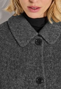 Close-up van een vrouw in een donkergrijze herringbone wollen jas met een brede kraag en zwarte knopen over een zwarte coltrui.