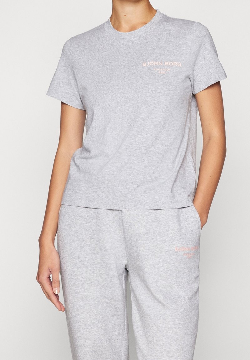 Kvinde iført en lys grå kortærmet T-shirt og matchende svedbukser med små pink "Björn Borg Stockholm 1956" logoer på brystet og låret.
