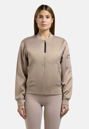 Frau mit einer beigen Satin-Bomberjacke mit gerippten Ärmelbündchen, Reißverschluss und Seitentaschen, die vor einem schlichten hellen Hintergrund steht.