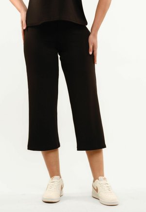 Persoon draagt zwarte cropped broek met wijde pijpen en witte sneakers met lichtbruine accenten, staand tegen een effen lichte achtergrond.