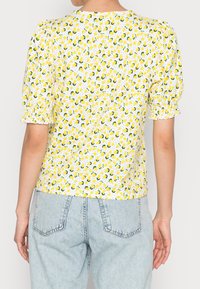 Kobieta w białej bluzce z krótkim rękawem z żółtym wzorem cytryn oraz bufiastymi rękawami, zestawiona z jasnoniebieskimi dżinsami z wysokim stanem.