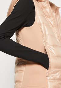 Kamizelka wypełniona w jasnoróżowym kolorze, z kieszenią na zamek błyskawiczny i smukłym fasonie. w zestawie z czarną bluzą z długim rękawem, ukazującą kontrastujące faktury.