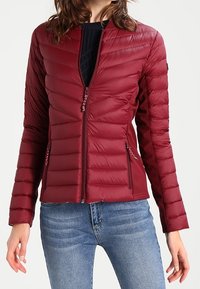 Maroon puffer jacket dengan desain quilted horizontal, kerah berdiri, ritsleting penuh, dan saku samping. Dipakai di atas atasan rajut navy.