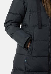 Giubbotto a cilindro nero con texture trapuntata, dotato di zip frontale, tasche laterali e un piccolo emblema blu e giallo vicino all'orlo.