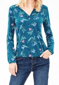Långärmad teal-topp med blommönster i lila och grönt, V-ringning, tillverkad av ett mjukt tyg. Bärs med blå jeans.