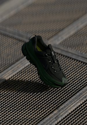 Black and green sneaker with a textured upper, cushioned sole, reflective accents, and elastic laces, set against a grid-like surface.