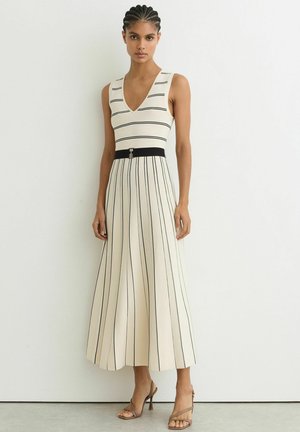 Femeie purtând o rochie crem fără mâneci cu dungi verticale negre, o curea neagră și sandale maro cu toc înalt, stând lângă un perete alb.