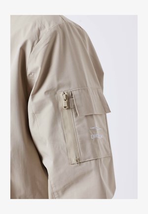 Beige jasje met een mouw die een rits- en klepzak heeft, met een klein geborduurd logo op de bovenarm.