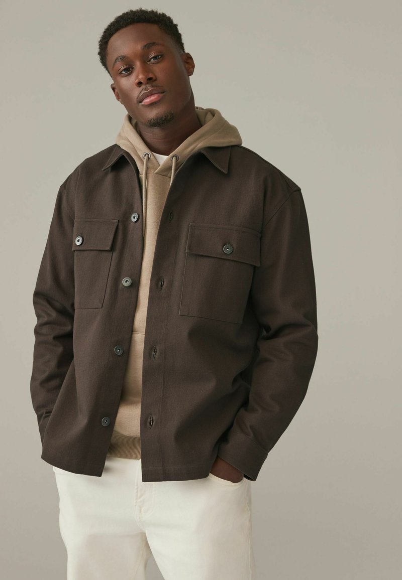 Brun knäppjacka med två bröstfickor, lager på lager över en beige hoodie. Vita jeans med avslappnad passform. Mjuk, texturerad tyg.