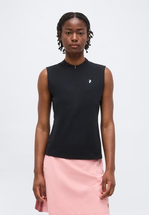 Kvinde med fletninger iført en ærmeløs sort top med et lille hvidt logo og et lyserødt skørt, stående foran en ensfarvet baggrund.