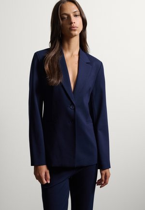 Donna che indossa un blazer blu scuro su misura e pantaloni abbinati, in piedi davanti a uno sfondo chiaro e semplice.