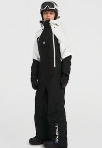 Traje de esquí en blanco y negro con bolsillos con cremallera, puños elásticos y un cuello alto. Incluye un logotipo en la pierna del pantalón y se combina con un casco.