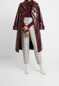 Burgunderfarbener, glänzender Trenchcoat über einem kastanienbraunen, gerippten Oberteil, kombiniert mit karierter, hochtaillierter Hose. Weiße Ankle Boots als Schuhe.