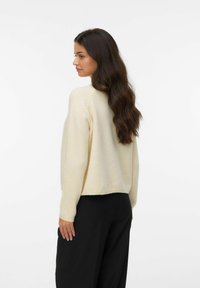 Pull en tricot beige avec une coupe décontractée et des manches longues. Doté d'un ourlet côtelé et d'une légère longueur courte, associé à un pantalon noir ample.