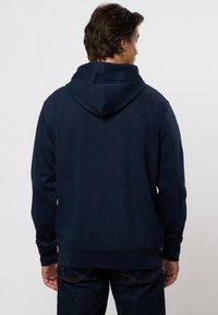 Hátrafelé néző férfi, sötét tengerészkék kapucnis pulóvert és sötét farmernadrágot visel, egyszínű világos háttér előtt.