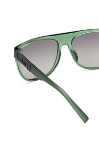 Occhiali da sole con tonalità verde, montatura trasparente e lenti grigio scuro sfumate. Presentano il marchio 'Timberland' sulla asta nera.