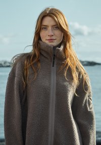 Fleece jakke i lysebrun med høj krave, lynlåslukning og tekstureret stof. Har et logo patch på venstre ærme.