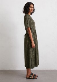 Dark green midi dress with a loose, short-sleeved design, featuring a tie waist and a subtle pattern. Worn with black sandals.