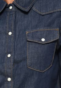 tmavě modrá džínová košile s vý pointed límečkem, předními knoflíkovými légy a jednou kapsou na náprsní. Kontrastní prošívání dodává látce detail.