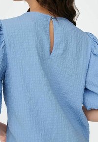 Blusa blu con texture, maniche corte e a sbuffo. Presenta un'apertura a chiave sul retro e una vestibilità leggermente ampia. Il materiale ha un motivo frastagliato.