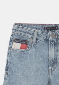 Světle modré denimové džíny s červenou, bílou a modrou nášivkou na přední kapse. Zahrnují stříbrný knoflík a štítek s nápisem "Tommy Hilfiger."