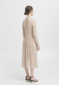 Ärmeloses, beiges Spitzenkleid mit tailliertem Schnitt, plissiertem Rock und Schlüssel-Ausschnitt hinten. Kombiniert mit schwarzen Riemchensandalen.