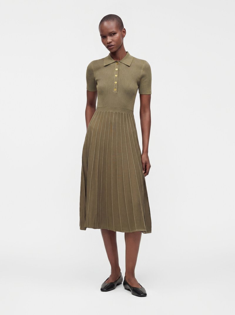 Femme portant une robe tricotée vert olive à manches courtes avec des boutons et une jupe plissée, debout les mains détendues le long du corps.