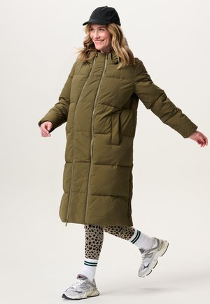 Eine Frau lächelt und schreitet voran, während sie einen langen olivgrünen Daunenmantel, Leggings mit Leopardenmuster, weiße Turnschuhe, gestreifte Socken und eine schwarze Mütze trägt.