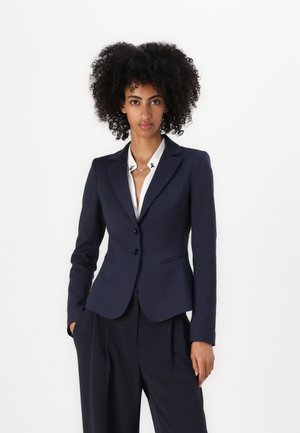 Marineblazer met een getailleerde silhouet, enkele knoopsluiting en ingelegde revers. gecombineerd met een wit overhemd met kraag, wat een verzorgde uitstraling geeft.
