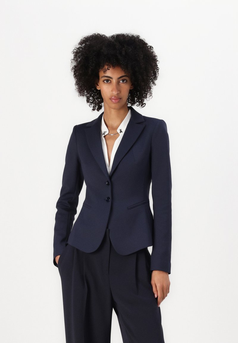 Marineblå blazer med en slim fit-silhuett, enkelt knappelukking og knappedetaljer. Kombinert med en hvit skjorte med krage, noe som gir et sofistikert utseende.