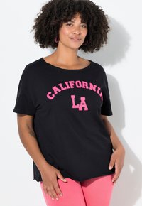 Mujer con cabello rizado, vestida con una camiseta negra de "California LA" y pantalones rosas, de pie contra una pared blanca con una sombra.