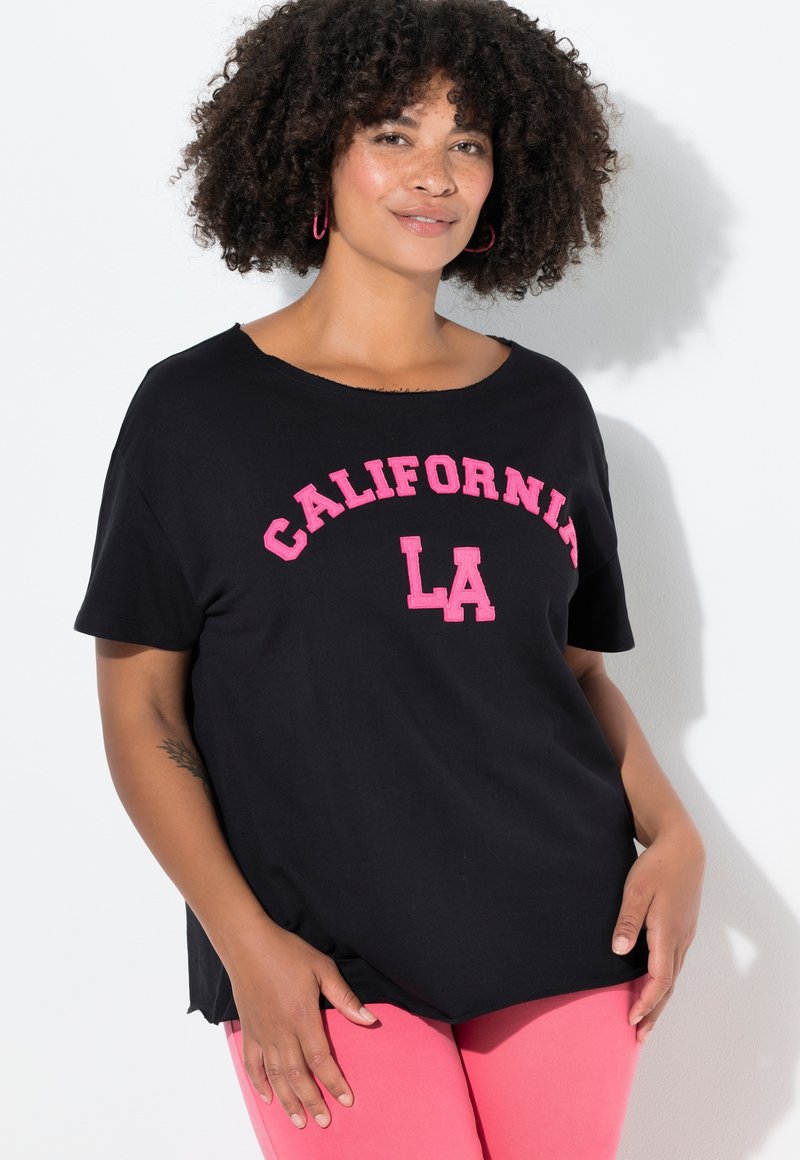 Mujer con cabello rizado, vestida con una camiseta negra de "California LA" y pantalones rosas, de pie contra una pared blanca con una sombra.